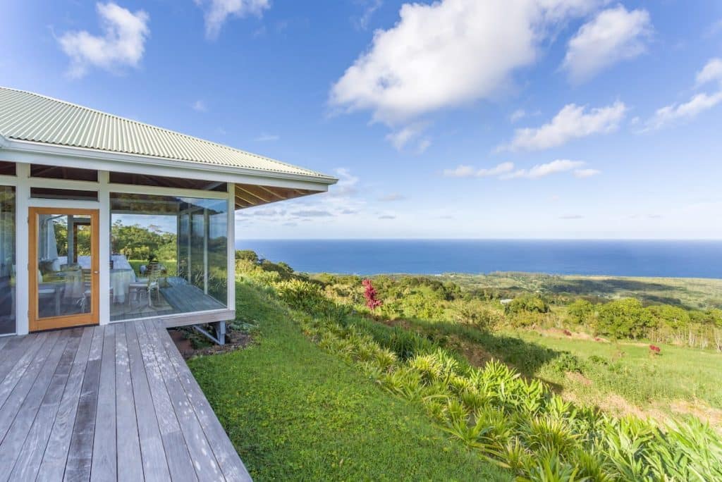 A modern house with large glass windows and a wooden deck overlooks a lush green landscape and ocean under a blue sky with scattered clouds.