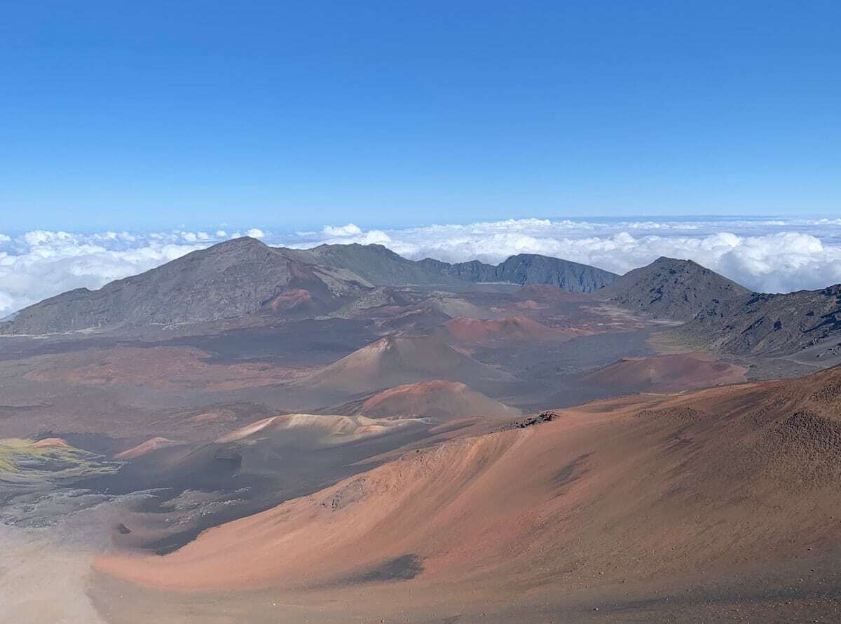 Panoramic view of a volcanic landscape with red and brown cinder cones, surrounded by mountains and a layer of clouds under a clear blue sky.