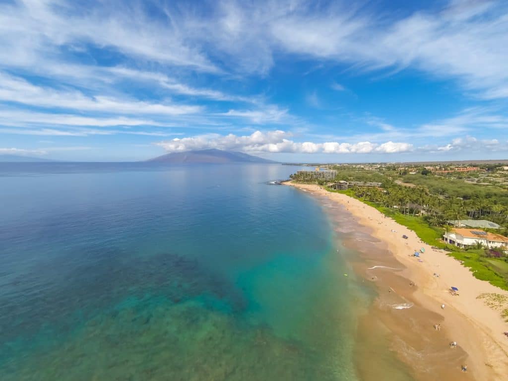 Aerial view of a sandy beach with turquoise water on a sunny day, featuring lush greenery and buildings along the shoreline, with a distant mountain visible under a partly cloudy sky.