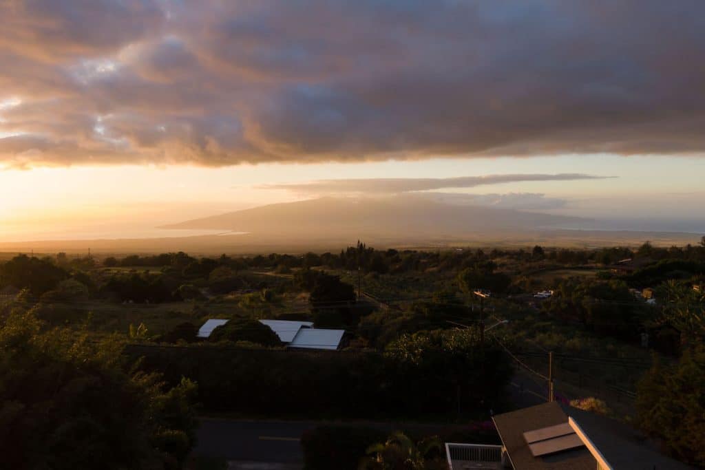 A scenic landscape at sunset with a mountain in the distance, partially covered by clouds, and lush greenery in the foreground.