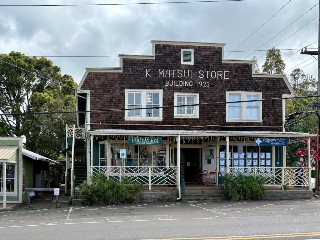 A historical building labeled 'K. Matsui Store, Building 1923' with a rustic wooden facade, featuring white-framed windows and a green and white porch, located beside a road with greenery and power lines.