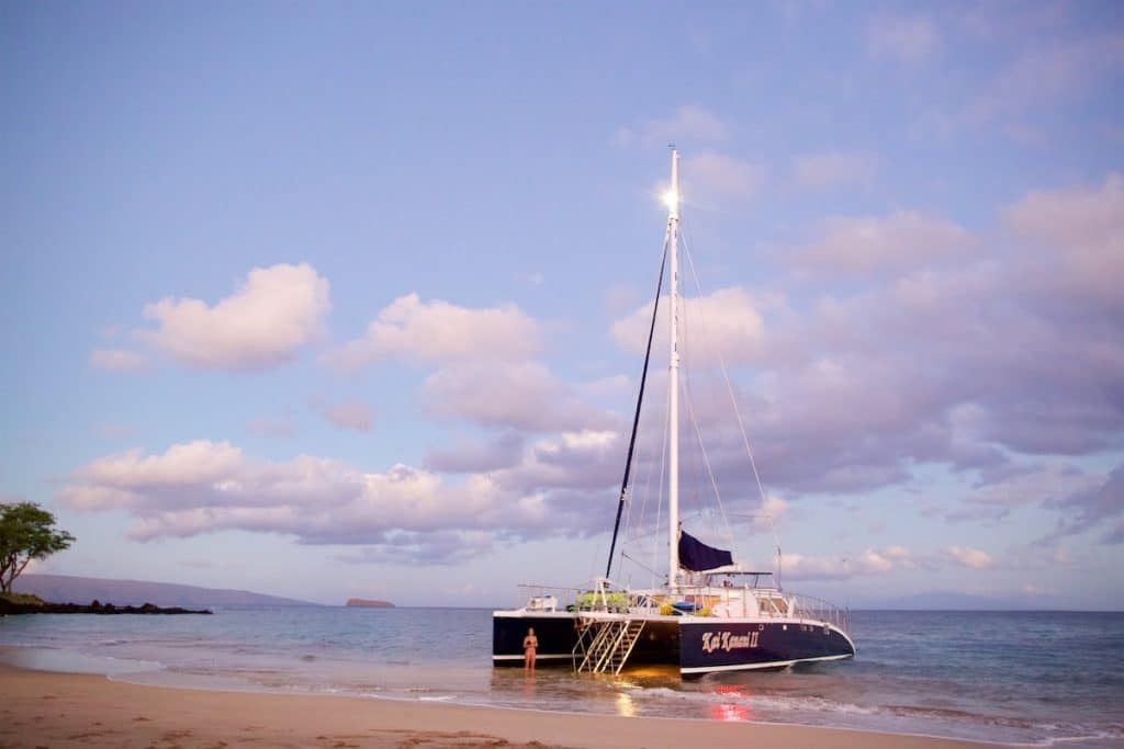 A catamaran with a tall mast is docked on a sandy beach under a cloudy sky at sunset, with a person standing in shallow water nearby.