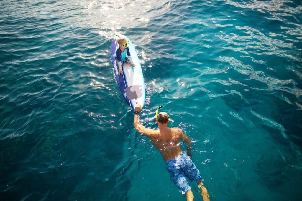 A child wearing a snorkel is sitting on a surfboard in the ocean, while an adult, also wearing a snorkel, swims next to the board in clear blue water.