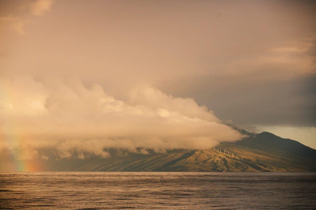 Landscape of a mountain partially covered by clouds with a faint rainbow on the left side, and wind turbines visible on the mountain slopes, set against an overcast sky.