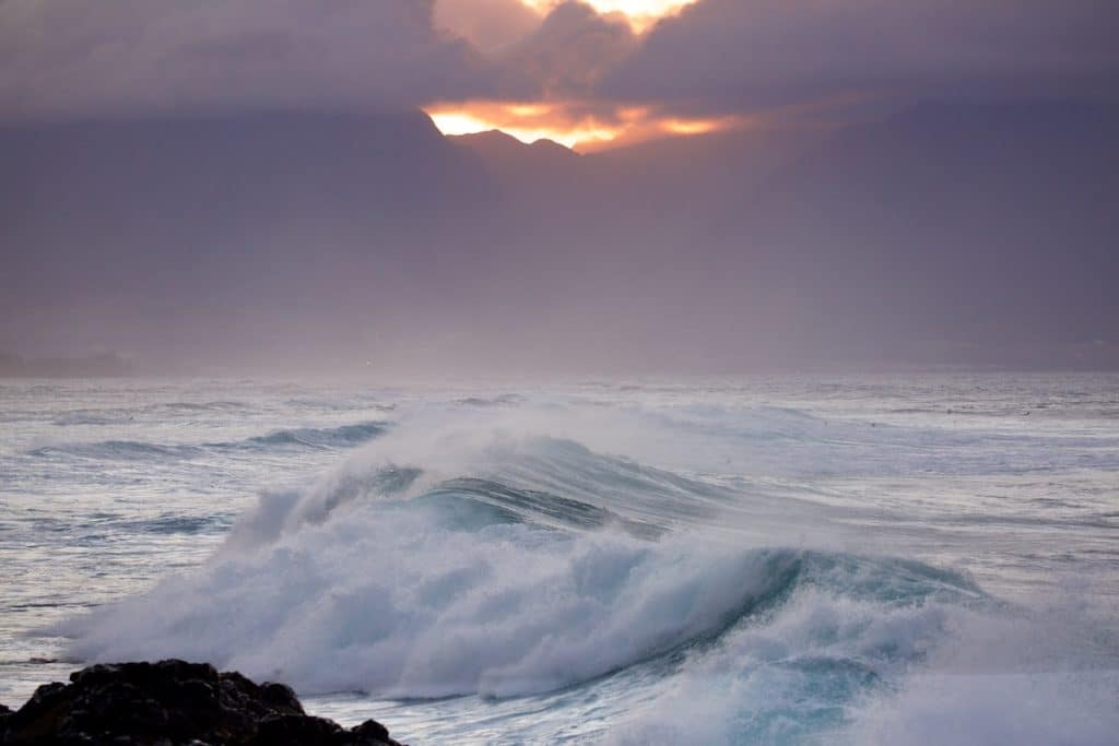 Waves crashing in the ocean with mist and mountains in the background, under a cloudy sky illuminated by the setting sun.