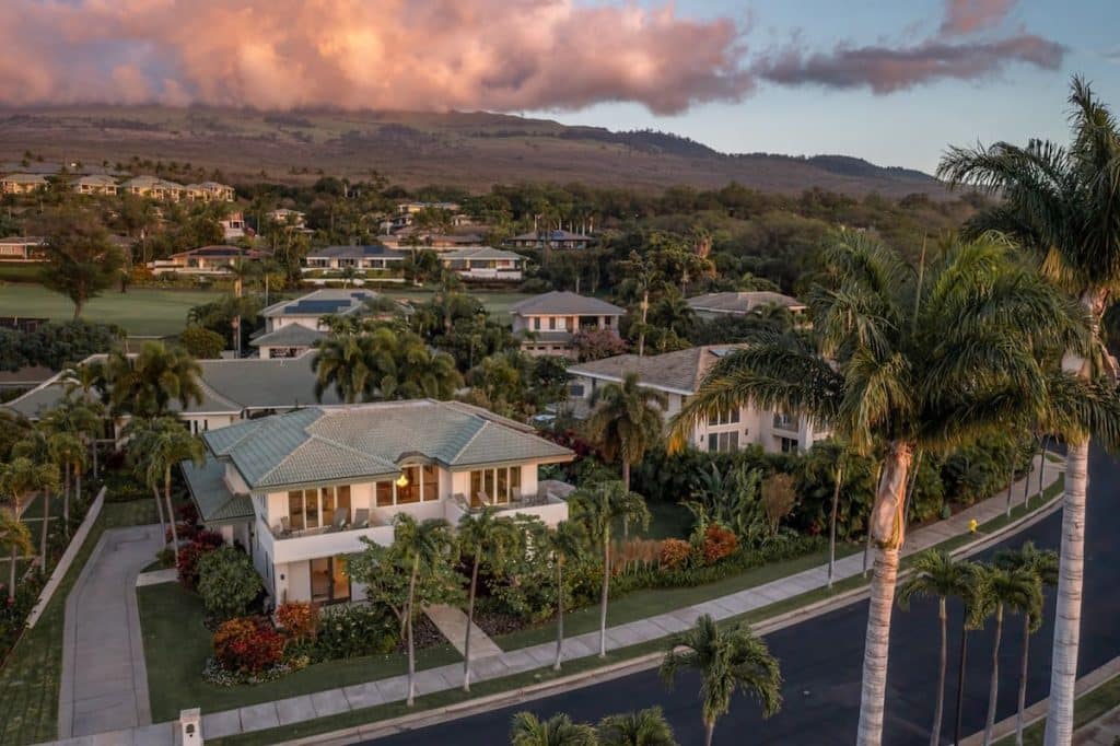 Aerial view of a suburban neighborhood with lush greenery, palm trees, and houses with well-maintained gardens under a sky with dramatic clouds at sunset.