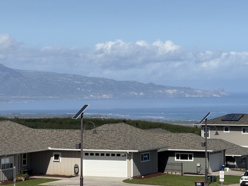 A scenic view of a residential neighborhood with green lawns and houses, overlooking a vast ocean and distant mountain range under a clear blue sky.