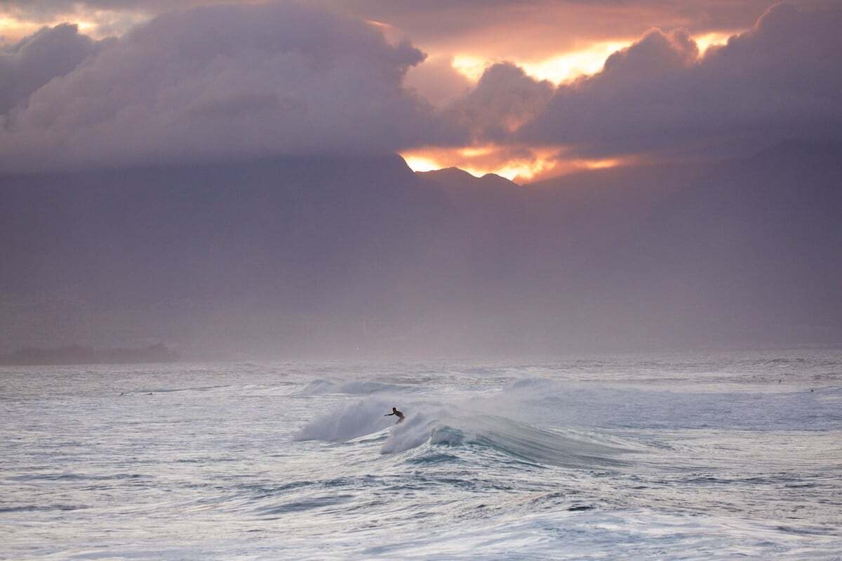 A surfer riding a wave in the ocean with a dramatic cloudy sky and mountains in the background at sunset.