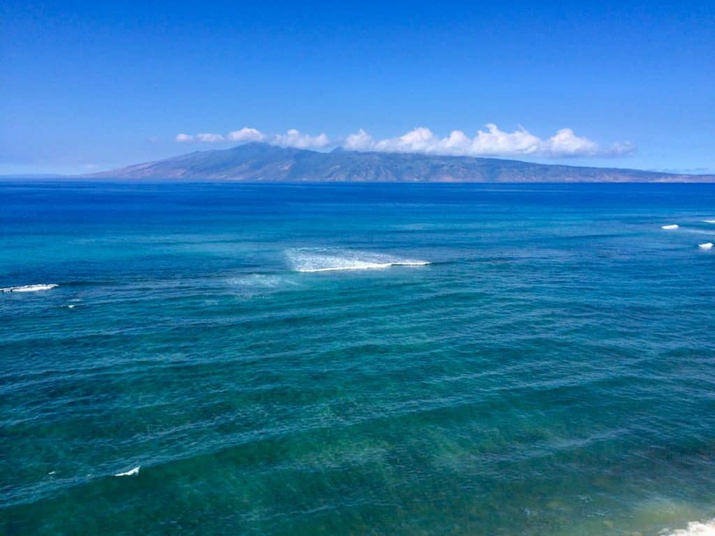 A vast expanse of blue ocean with gentle waves, under a clear blue sky, and an island with mountainous terrain visible in the distance.