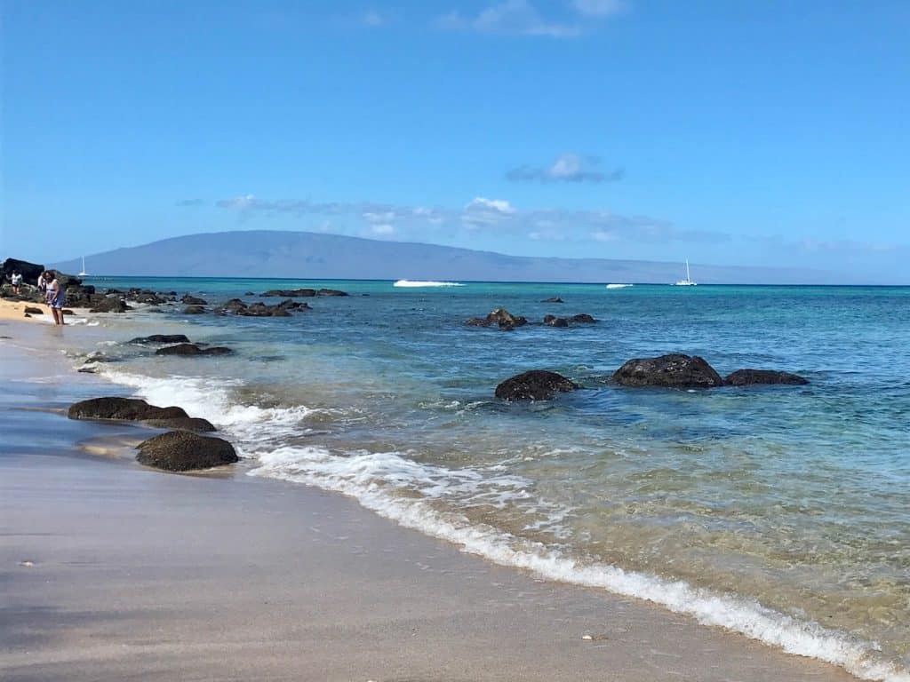 A sandy beach with scattered rocks along the shore, clear blue water, and a distant sailboat on the horizon under a clear sky.