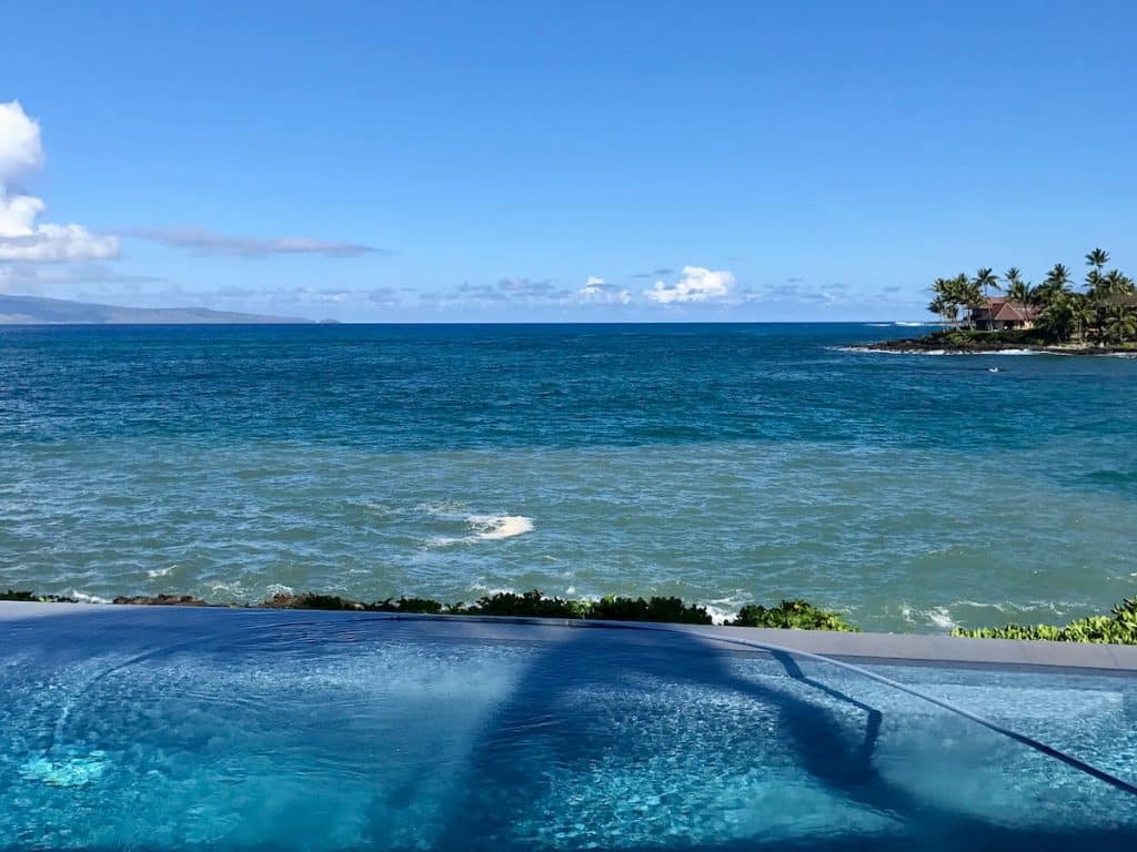An infinity pool overlooking a vibrant blue ocean with a small peninsula featuring palm trees and a building in the background under a clear blue sky.